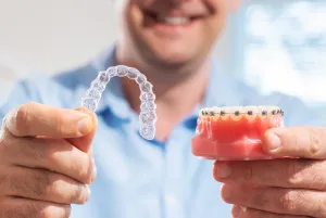 a man holds a clear aligner next to a model with braces